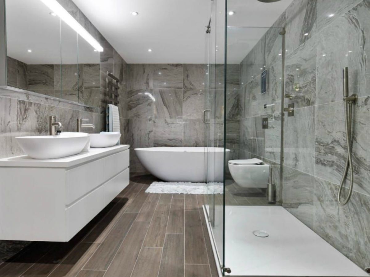 Grande salle de bain marbre veiné gris, sol imitation parquet brun, baignoire îlot et douche vitrée avec pommeau chrome.