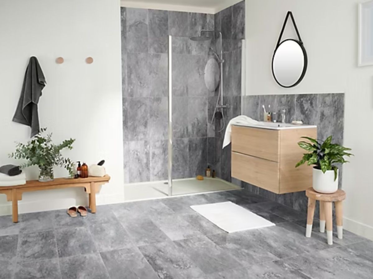Salle de bain carrelage gris pierre au sol et paroi douche, meuble chêne, banc rangement et miroir à lanière cuir.