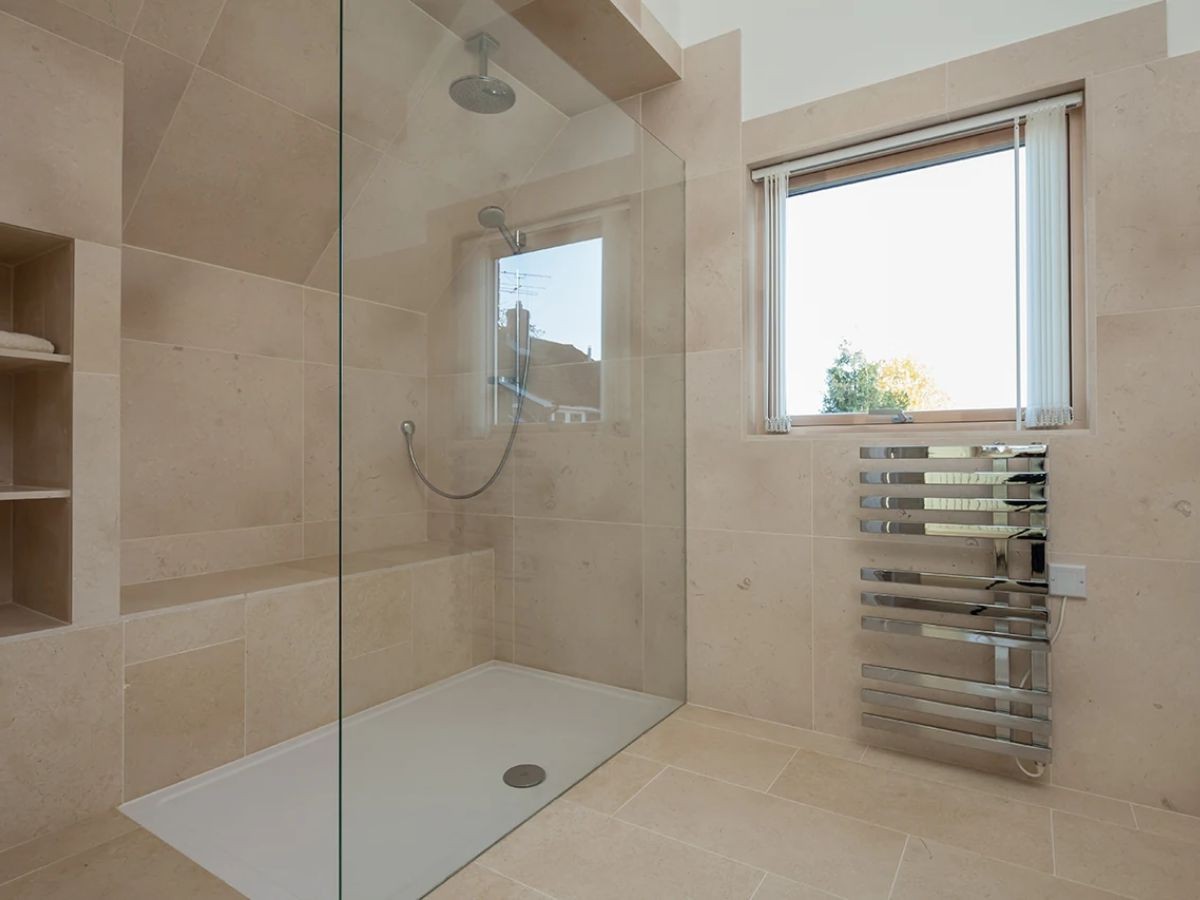 Douche spacieuse carrelée beige pierre avec banc intégré, pommeau plafond, niche verticale et sèche-serviettes chrome.
